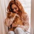 Uma mulher ruiva sorridente abraça carinhosamente um gato persa laranja fofo perto de uma janela com cortinas brancas.